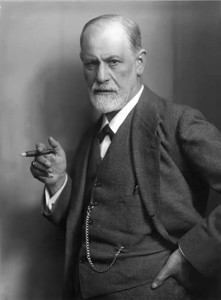 Black and white portrait of Sigmund Freud holding a cigar, posing with a serious expression, wearing a dark suit, captured in a classic early 20th-century style.