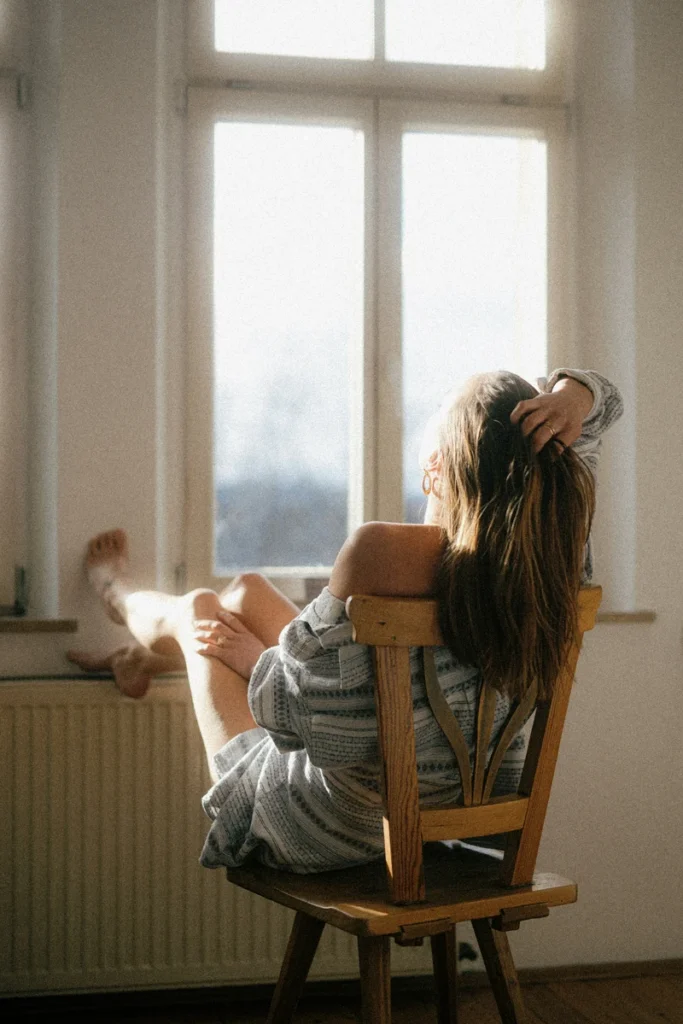 leo rising confidence woman sitting with her back to the camera by a window, feet resting against the wall, expressing self-assurance, presence, and quiet strength