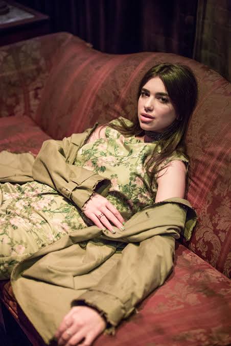 Dua Lipa birth chart image showing Dua Lipa sitting on a sofa, capturing her relaxed, elegant presence and modern pop star style.