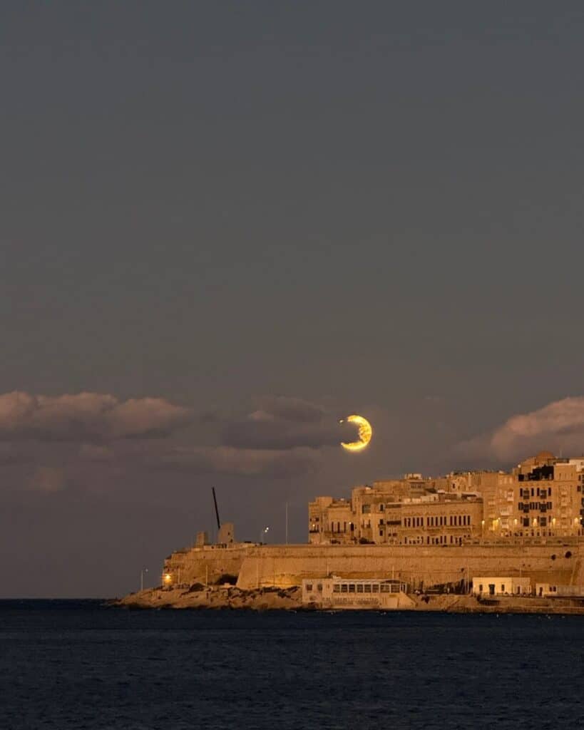 Solar eclipse in Leo glowing above a coastal city at dusk, with dark clouds, illuminated historic buildings, and the sea in the foreground.