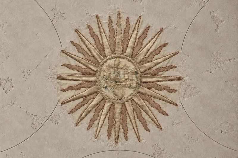 An antique embroidered sun motif on a textured neutral wall, symbolising illumination and self knowledge through an online astrology reading.