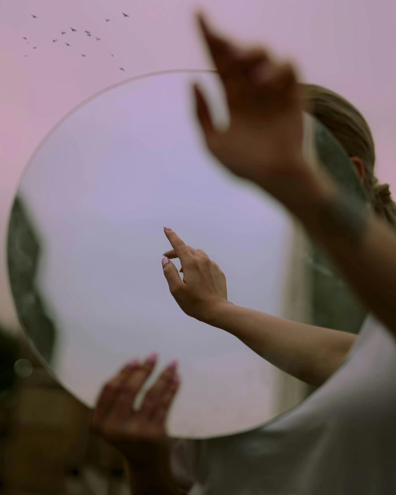 Mirror in pink and purple tones reflecting a pair of hands, evoking self-awareness, introspection, and the birth chart as a mirror of the soul.