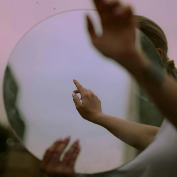 Mirror in pink and purple tones reflecting a pair of hands, evoking self-awareness, introspection, and the birth chart as a mirror of the soul.